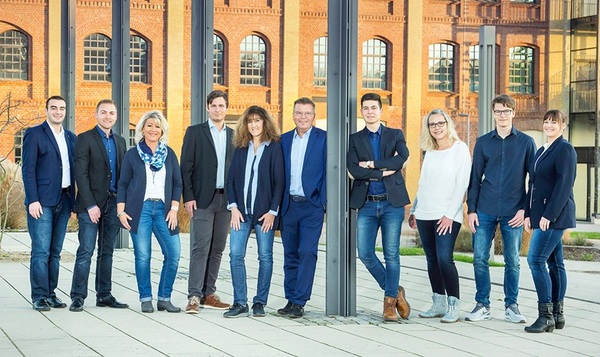 Gruppe von Mitarbeitern steht vor moderner Backsteinfassade, Teamfoto einer Firma draußen.